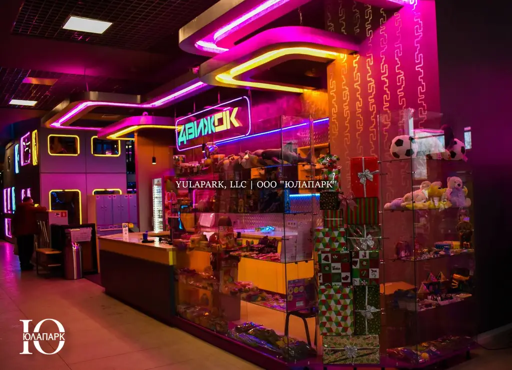 Вход и входная группа в детский активити-парк Движок в Омске с неоновой розовой, синей и желтой подсветками, большим ресепшеном и витриной с подарками и сладостями.