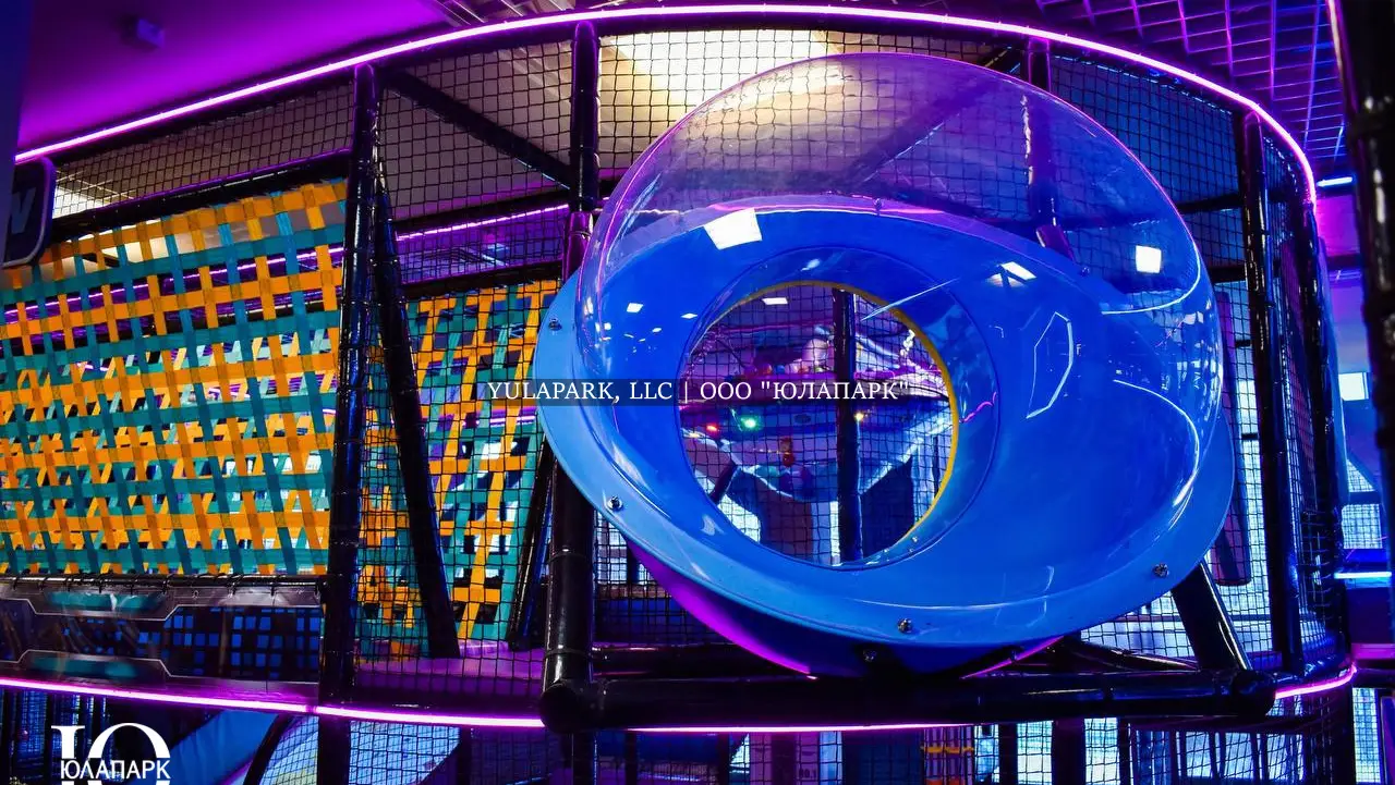 Прозрачная сфера в виде ока в центре детского лабиринта в активити-парке с вязанным мостом на заднем фоне.