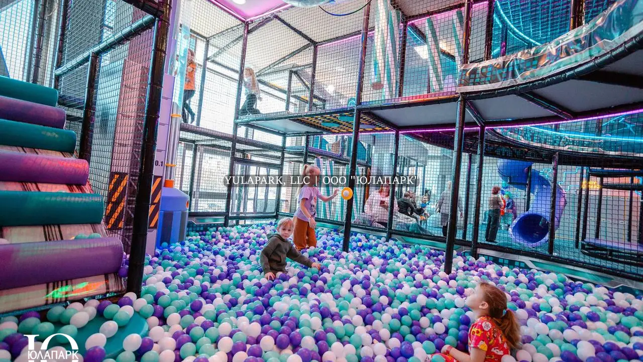 Дети играют в сухом бассейне с фиолетовыми, голубыми и белыми шариками на фоне большого высокого лабиринта с неоновой подсветкой и множеством горок в активити-парке в Омске.