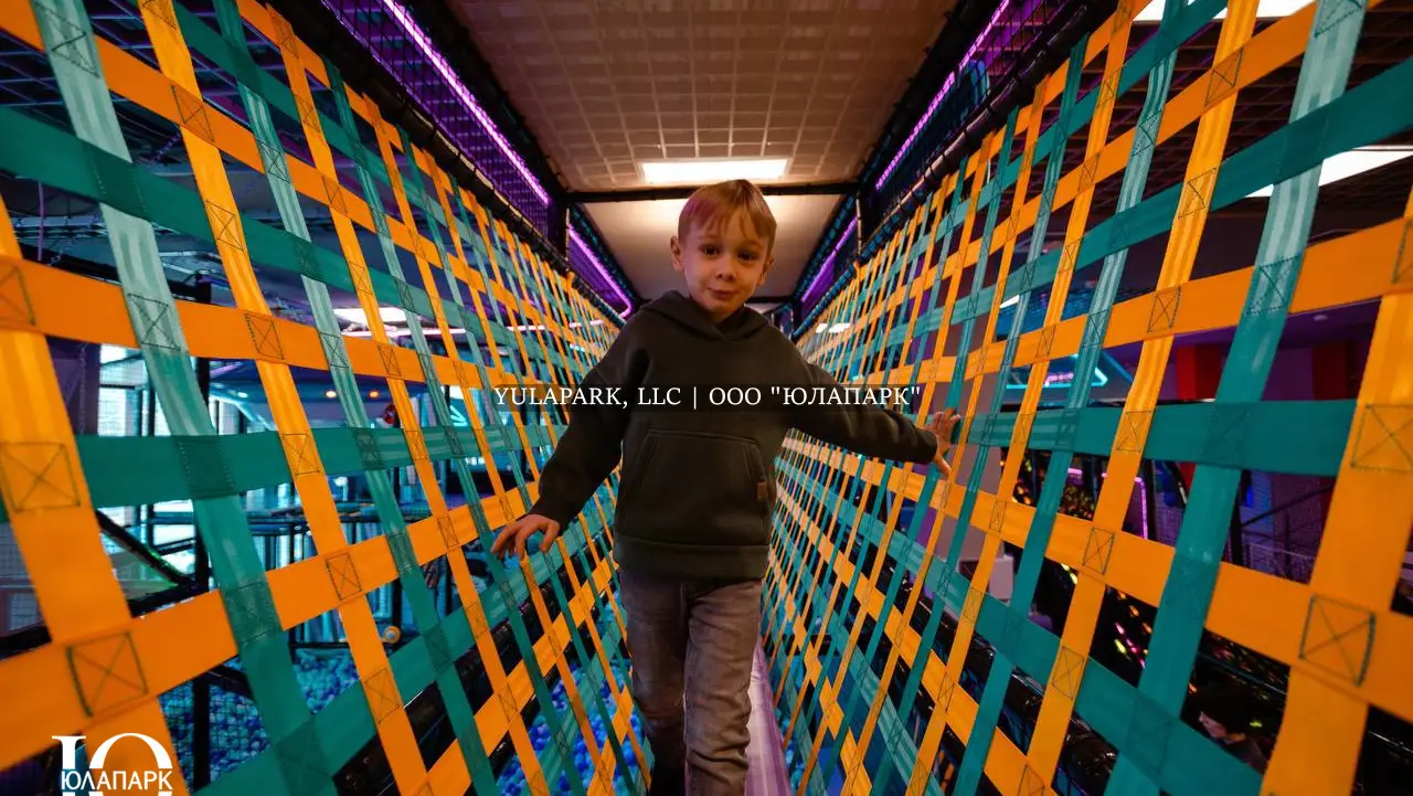 Вязанный мост с синими и оранжевым плетением или полосками, по которому идет мальчик, ребенок в парке Движок.