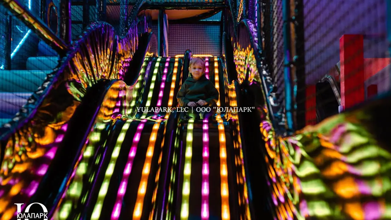 Черная пластиковая горка с неоновой анимированной подсветкой в виде дождика на фоне большого лабиринта, откуда съезжает мальчик в парке Движок.