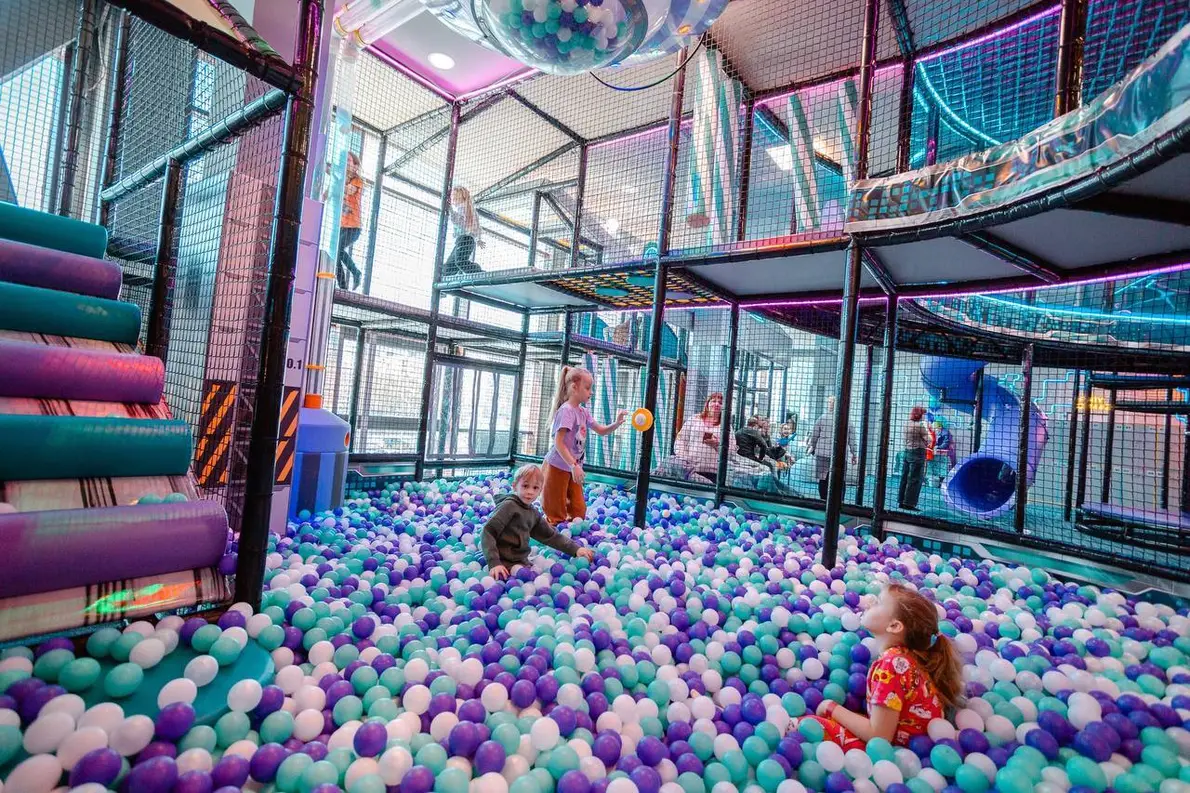 Дети играют в сухом бассейне с фиолетовыми, голубыми и белыми шариками на фоне большого высокого лабиринта с неоновой подсветкой и множеством горок в активити-парке в Омске.