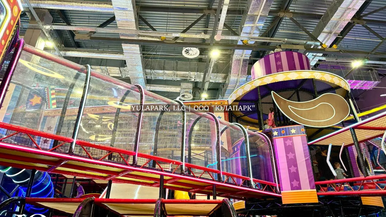 Длинный прозрачный мост с неоновой подсветкой и красной башней в виде купола цирка в детском развлекательном парке Флик Фляк.