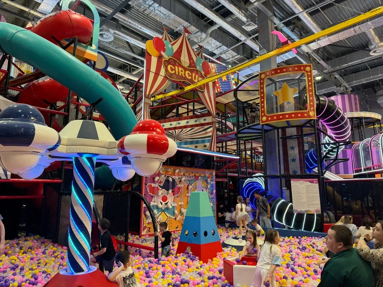 Большая игровая зона в виде цирка для детей с большим сухим бассейном, различными каруселями, горками, скайрайдером и лабиринтами с неоновой подсветкой в детском развлекательном парке Флик-Фляк.